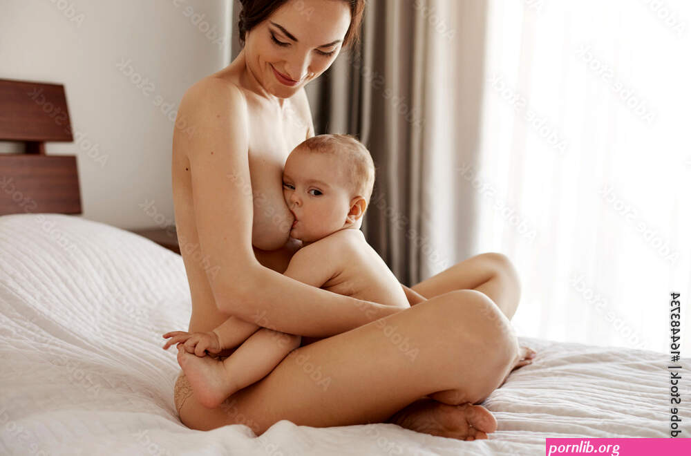 Young attractive nude mom breastfeeding hugging her newborn baby smiling sitting on bed at home. Stock Photo