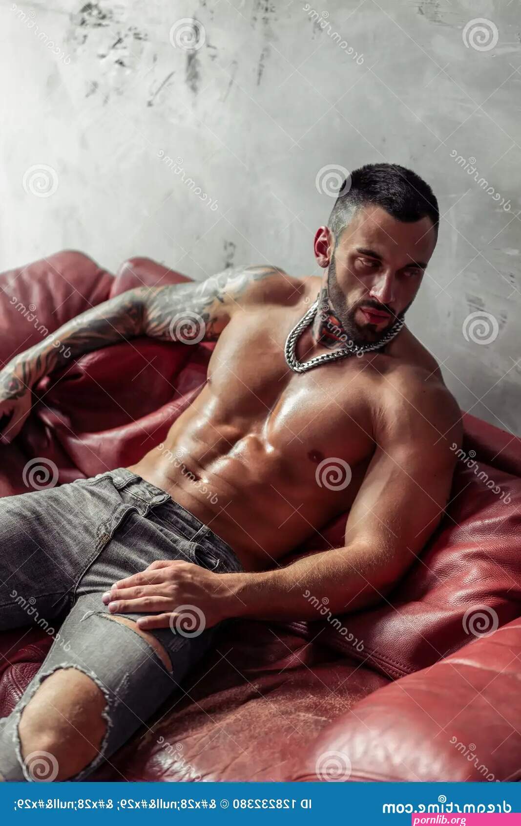 Fashion Portrait of Naked Male Model with Tattoo and a Black Beard Sitting in Hot Pose on Red Leather Sofa. Loft Room Interio Stock Photo