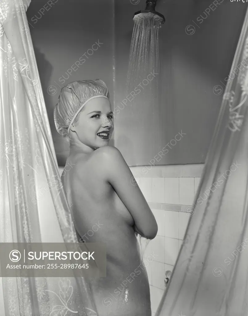 Vintage Photograph. Nude woman wearing only shower cap standing in shower with water running viewed from behind with head turned smiling